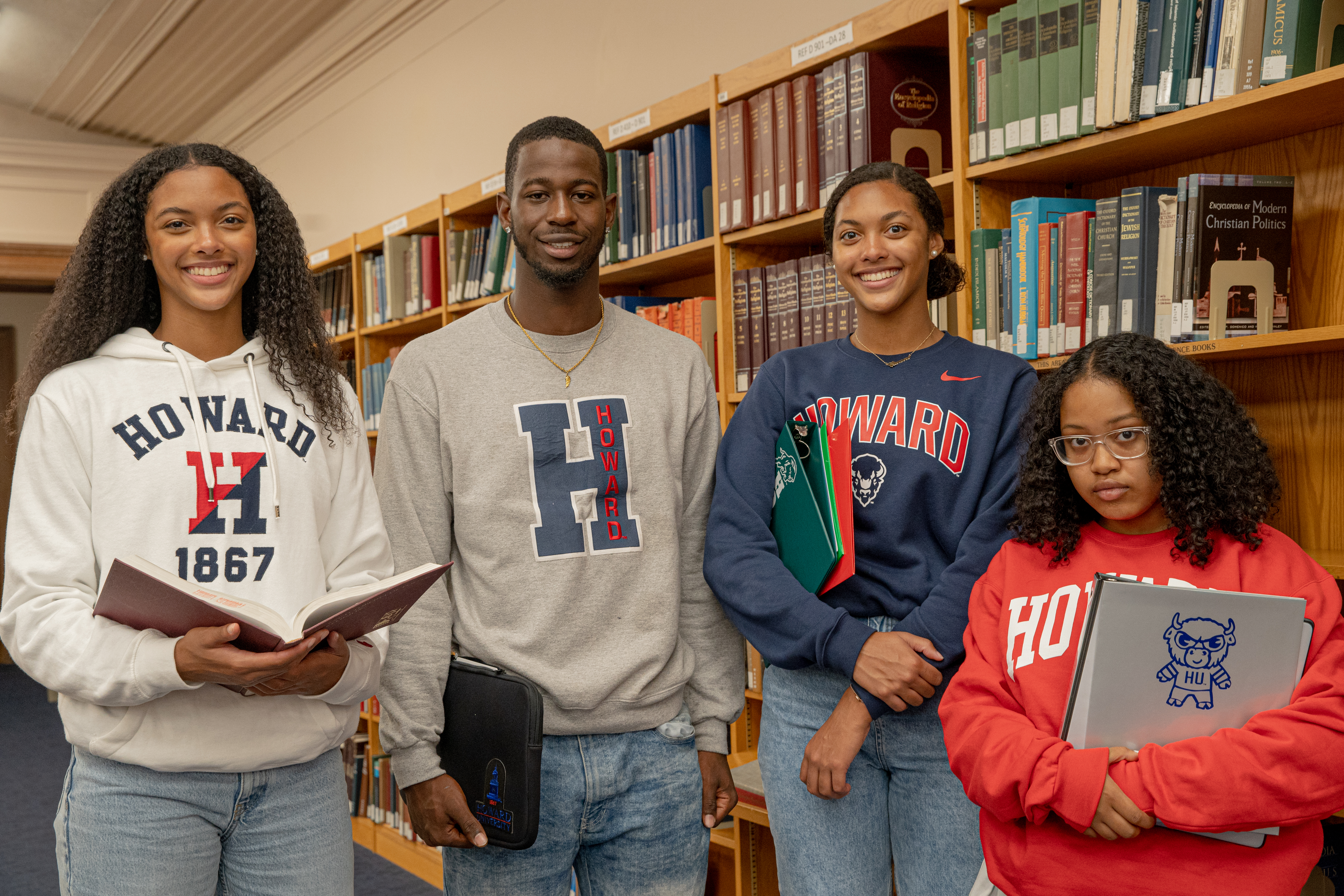 Four students in Howard apparel holding books in the library